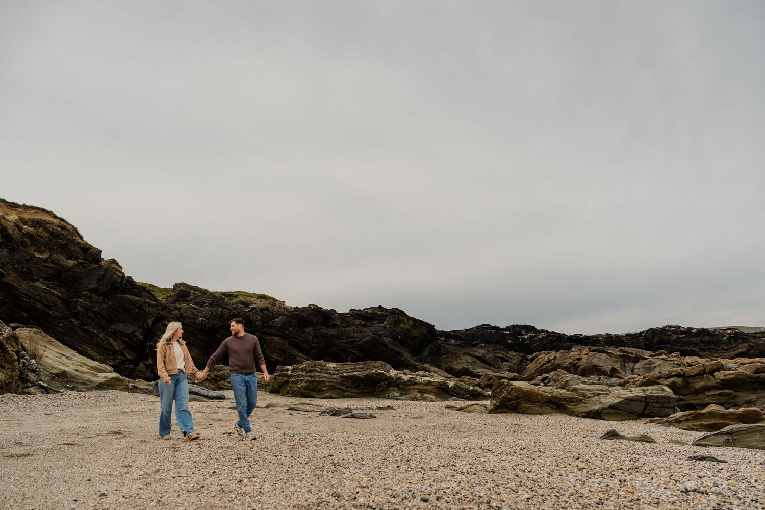 Couple holding hands walking on a rocky pebble beach with dark cliffs and overcast sky.