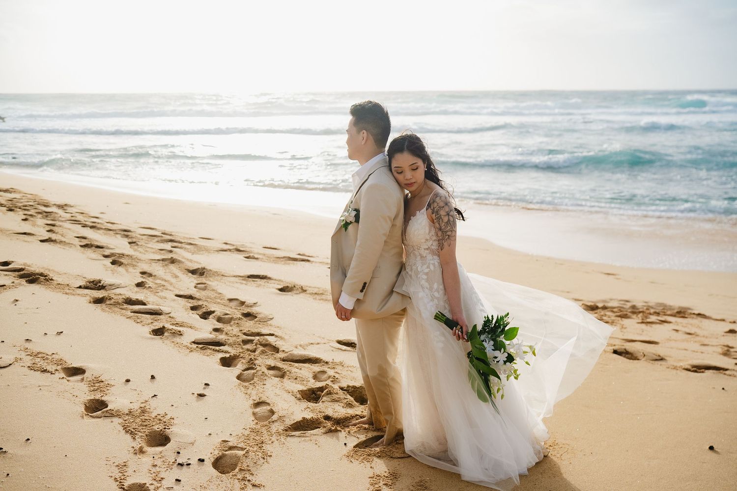 Bride and groom standing back-to-back on a sandy beach, bride holding tropical bouquet with ocean waves behind them.