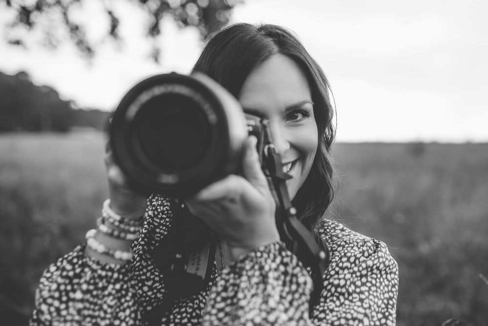 Photographer smiling behind professional camera lens in outdoor setting.
