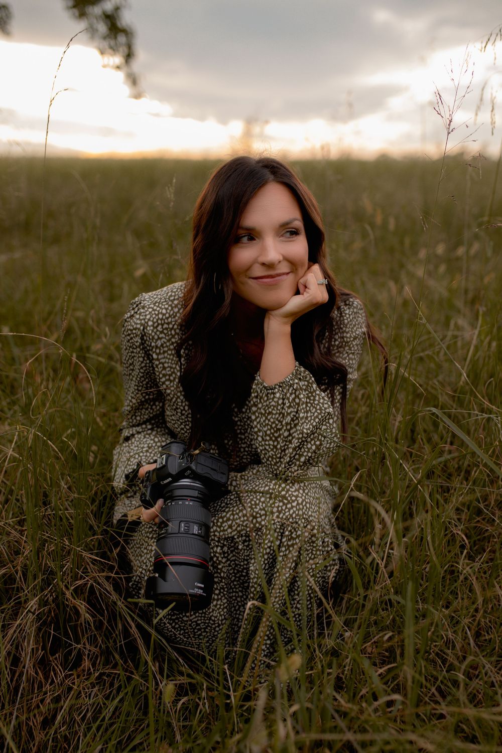 A photographer in a floral dress sits in a grassy field at sunset with a professional camera.