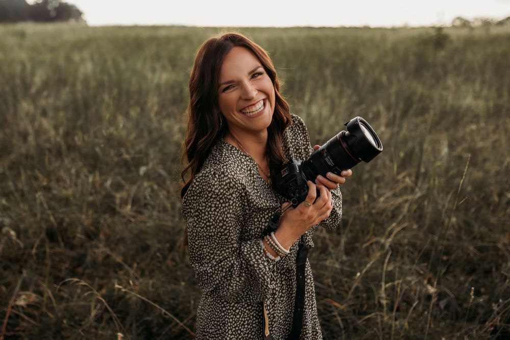 Professional photographer in a leopard print top captures outdoor portraits in a golden field at sunset.