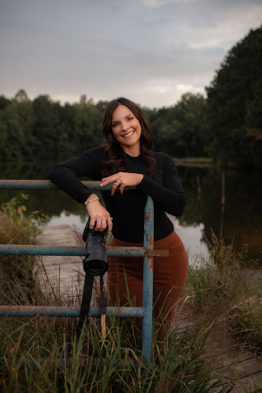 A person in black clothing poses near a lake at sunset with a rustic blue railing in the foreground.