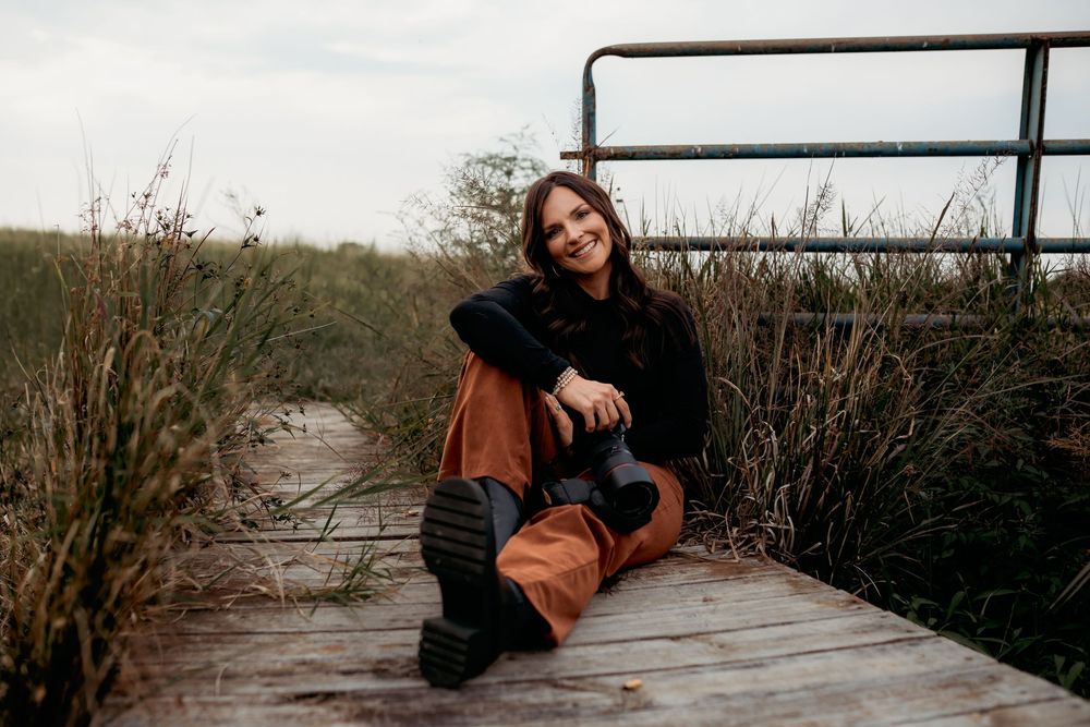 A person in a brown jacket sits on a wooden boardwalk surrounded by coastal vegetation at sunset.