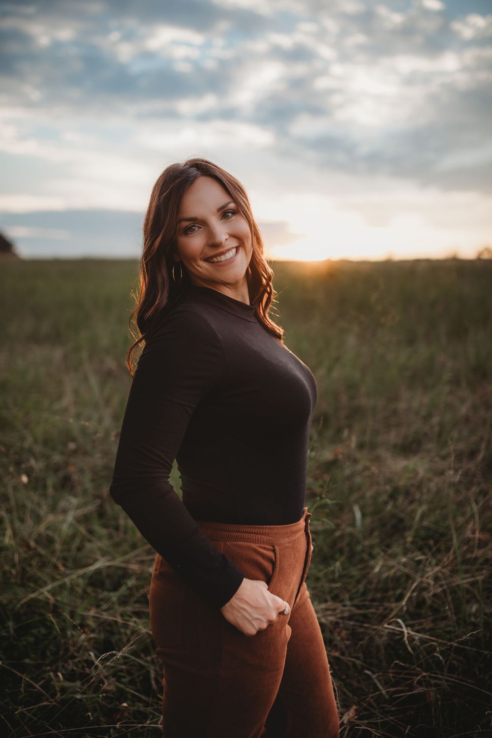 Woman in dark sweater poses in a field during golden hour sunset with dramatic cloudy sky.