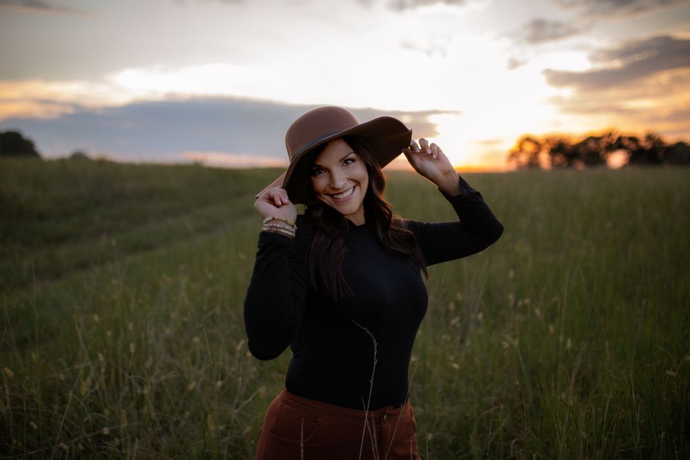 Person in a wide-brimmed hat stands in a grassy field during golden hour sunset.