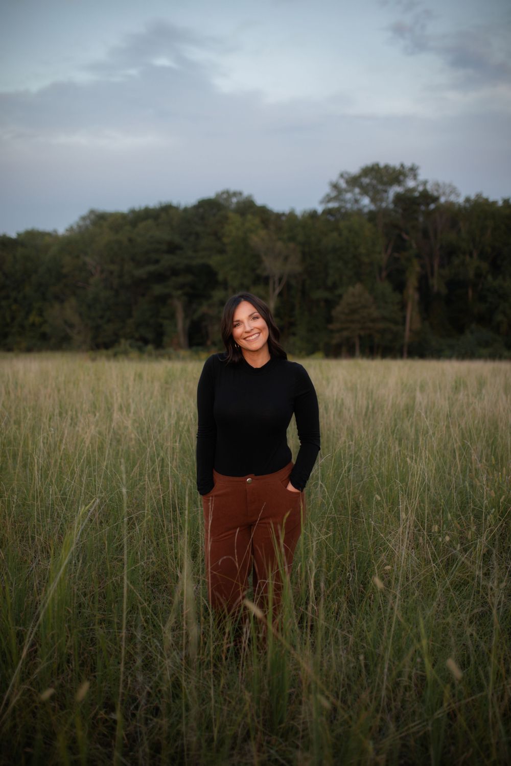A person wearing a black top and brown pants stands in a tall grass field during an overcast day.
