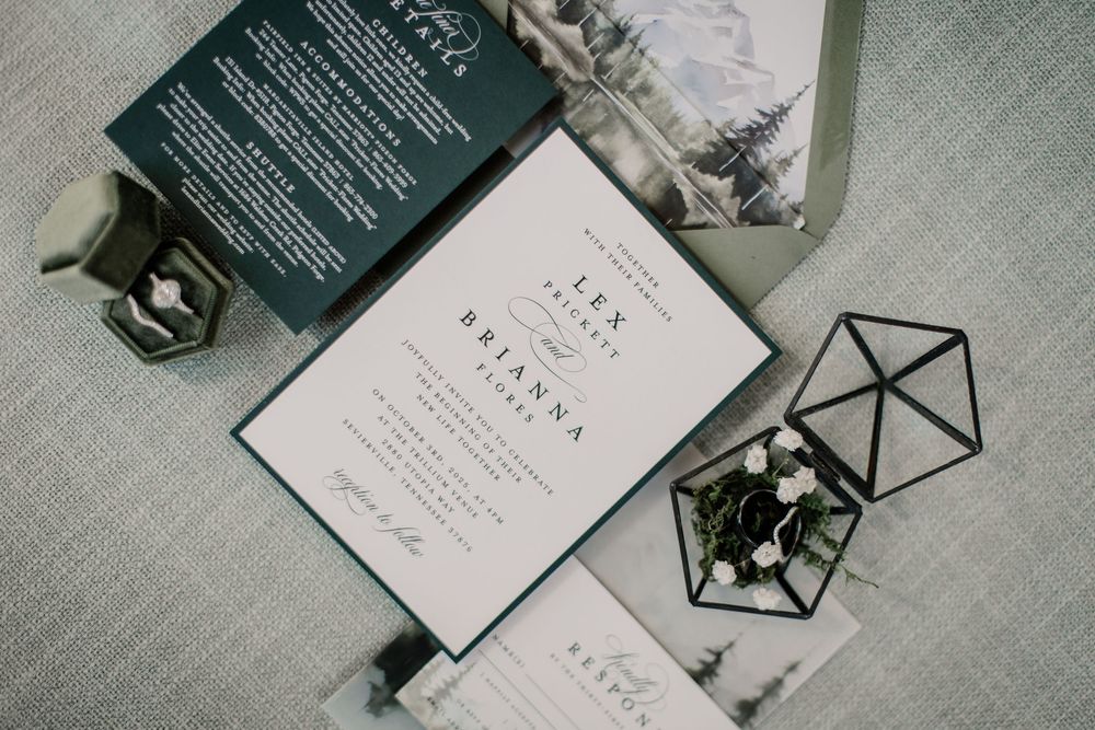 Elegant wedding invitation suite with sage green accents, geometric ring box, and mountain-themed stationery on gray linen.