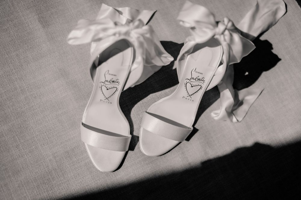 White bridal heels with bow ankle straps and heart detail on sole, photographed in black and white.