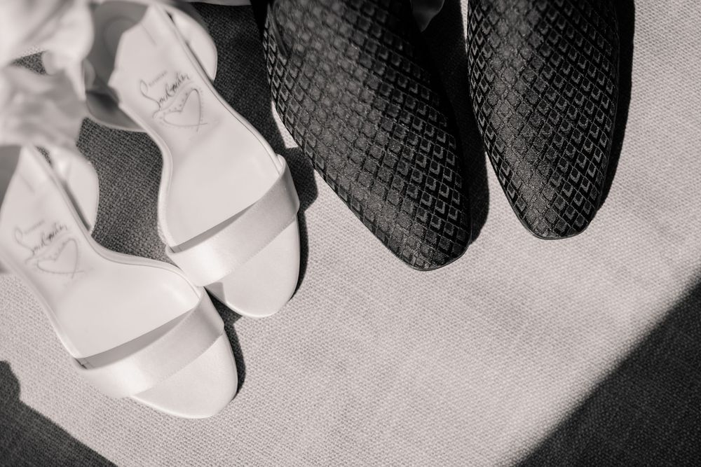 Black and white photo of elegant white bridal heels beside dark groom dress shoes on a textured surface.