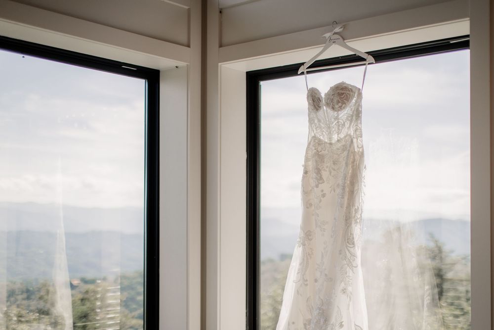 Strapless lace wedding gown hanging in window with mountain landscape view on wedding day.