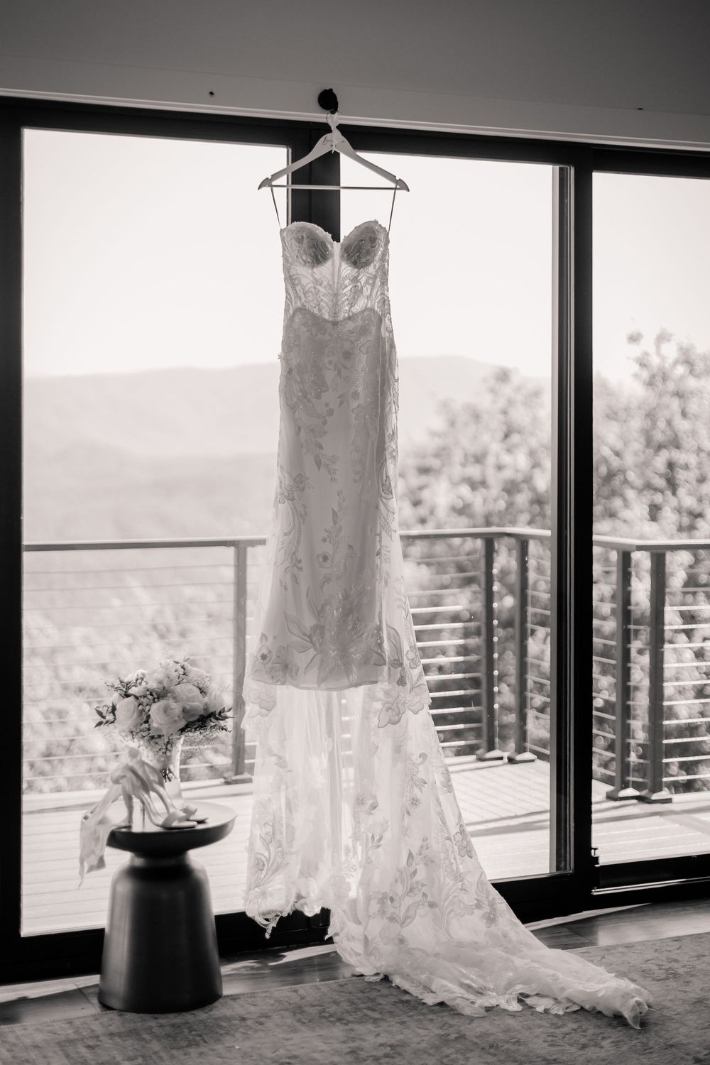 Black and white photo of a lace wedding gown hanging by a window with mountain views.