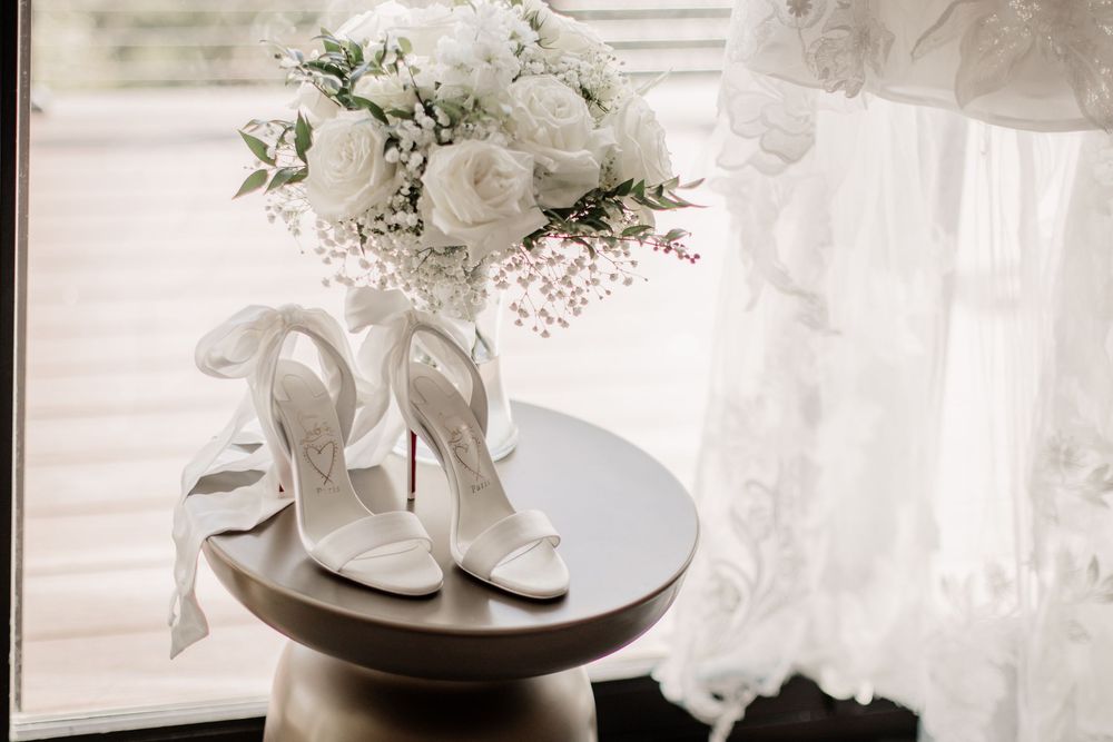 White bridal heels with ribbon ties displayed on a pedestal table beside a white rose bouquet.