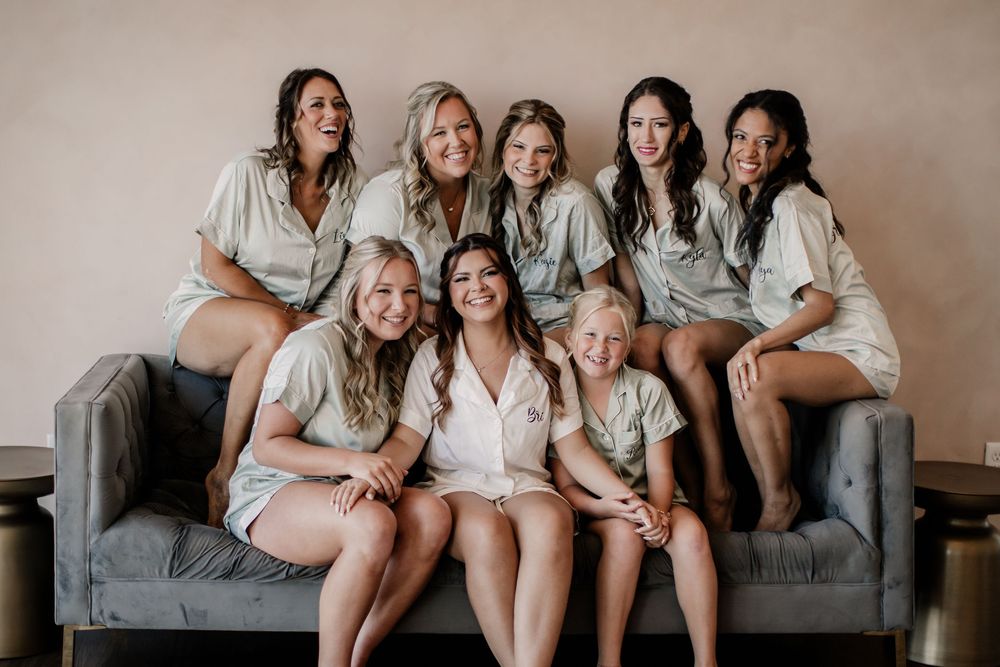 Bride and bridesmaids smiling together in matching satin robes seated on a gray couch.