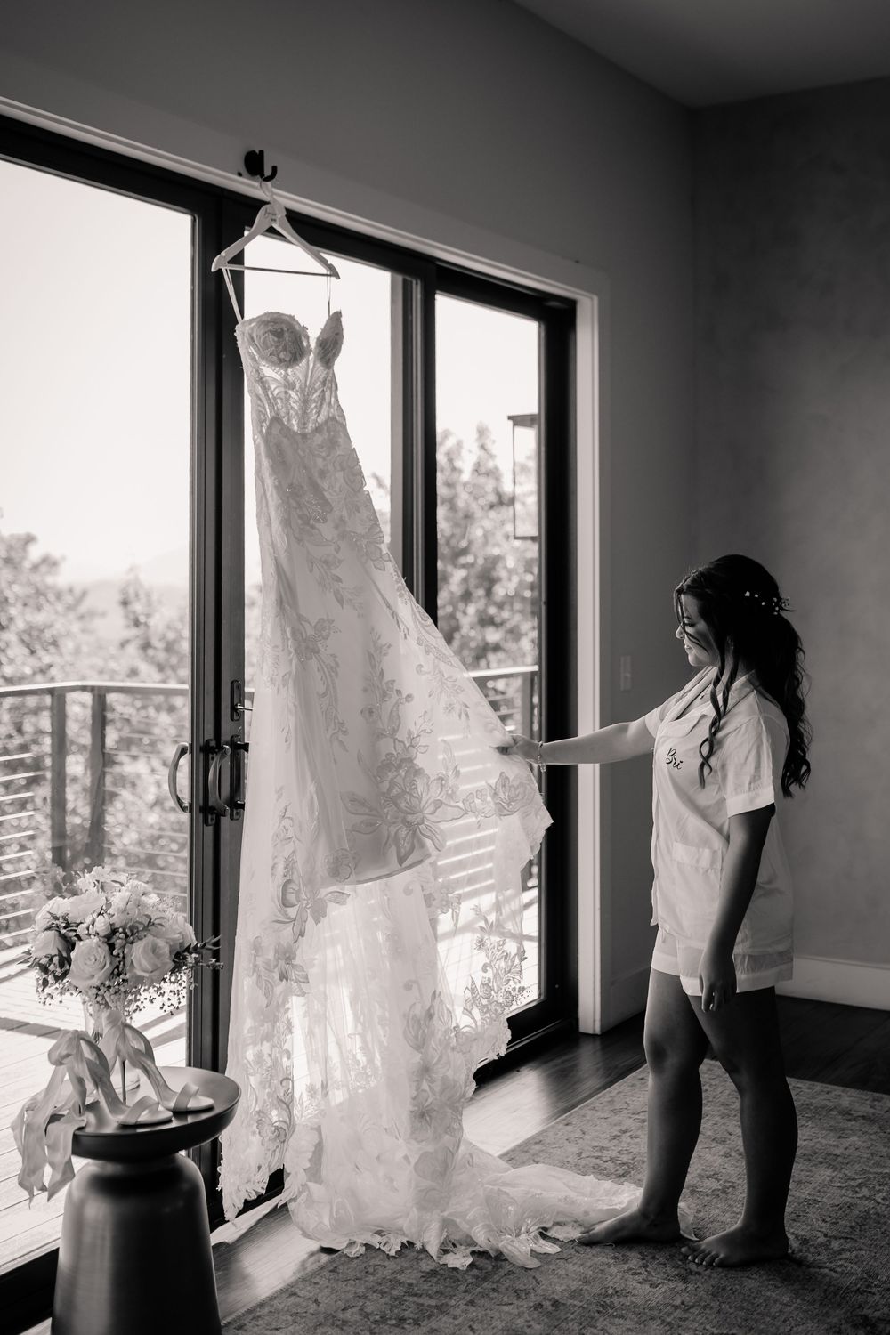Bride in robe admiring her lace wedding gown hanging by a window in black and white.