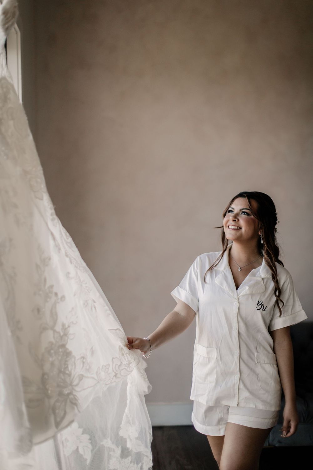 Smiling bride in white pajamas admiring her lace wedding dress hanging in a softly lit room.