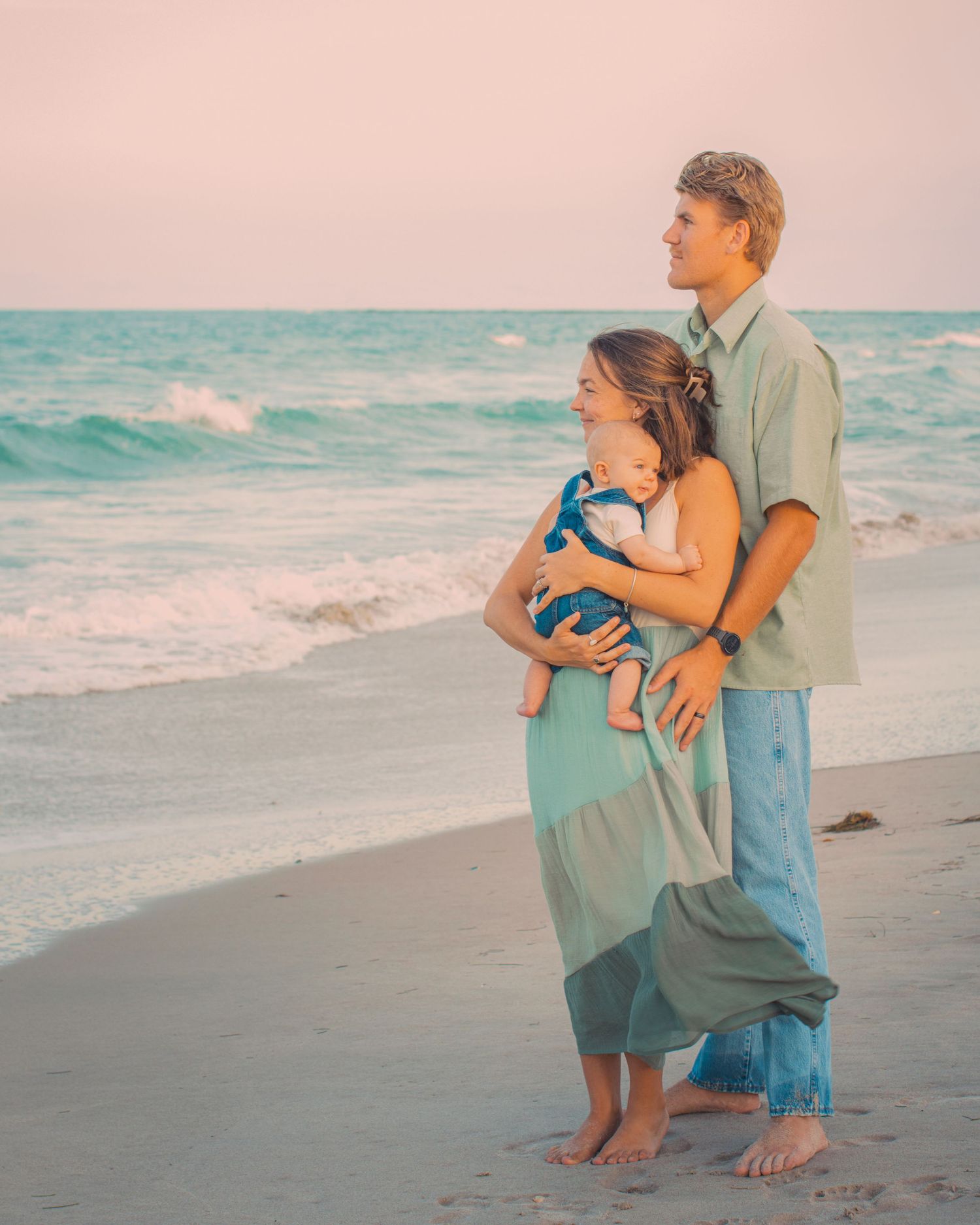 Young couple holding baby on beach at sunset, wearing matching green outfits, waves and pastel sky in background.