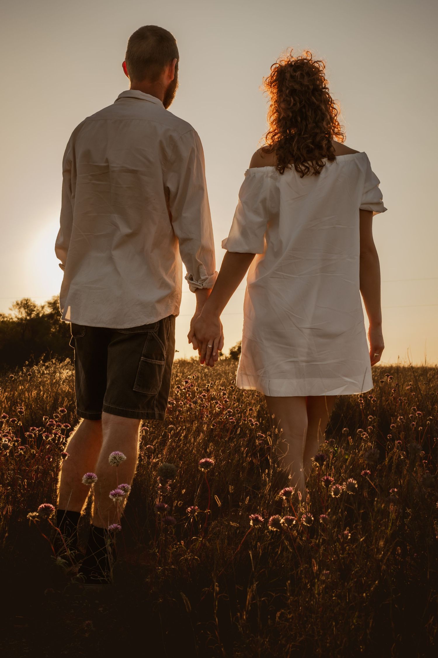 Couple holding hands in a golden sunset field, both wearing white, viewed from behind among wildflowers.