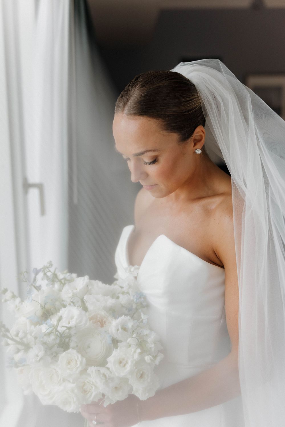 Elegant bride in a strapless white gown and veil gazes down at her white floral bouquet by a bright window.