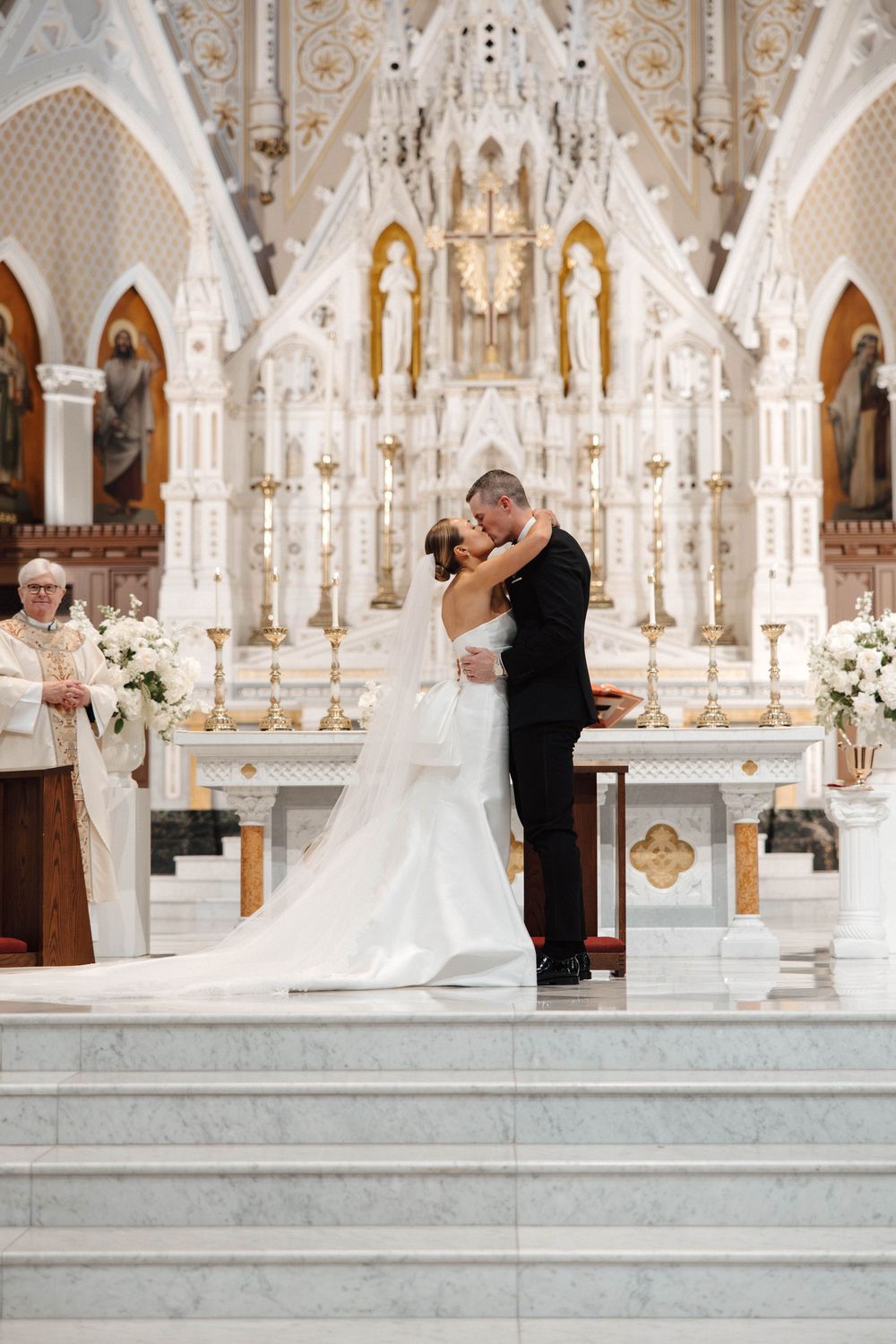 Bride and groom share first kiss at ornate white church altar during wedding ceremony.