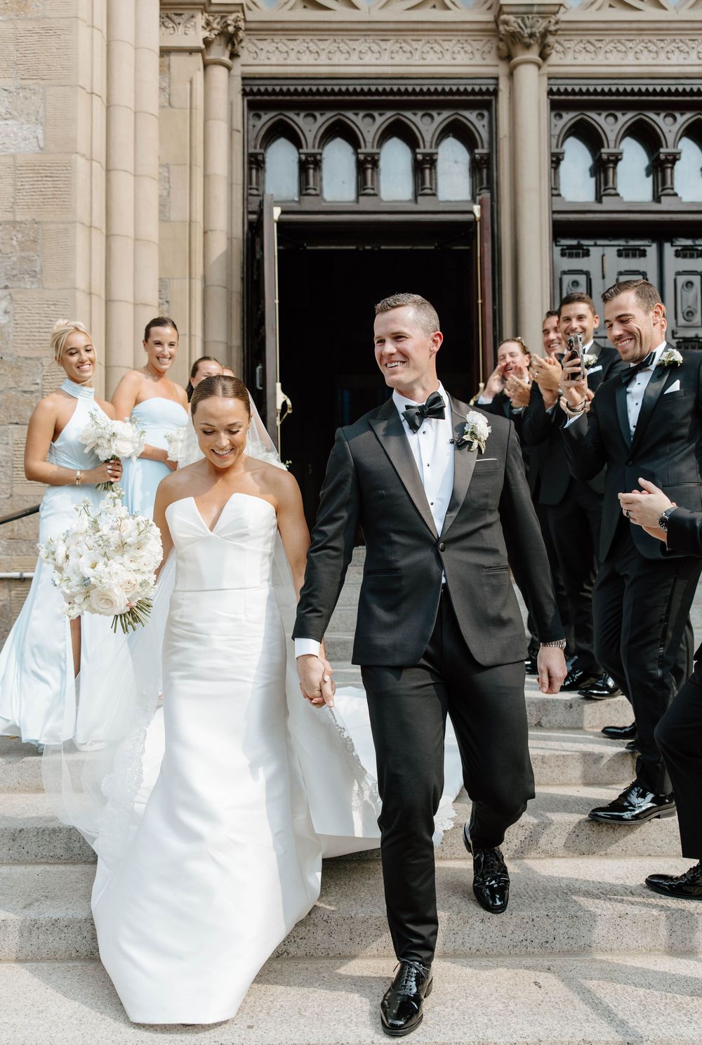 Newlyweds exit grand Gothic church entrance as wedding party celebrates behind them with champagne glasses raised.