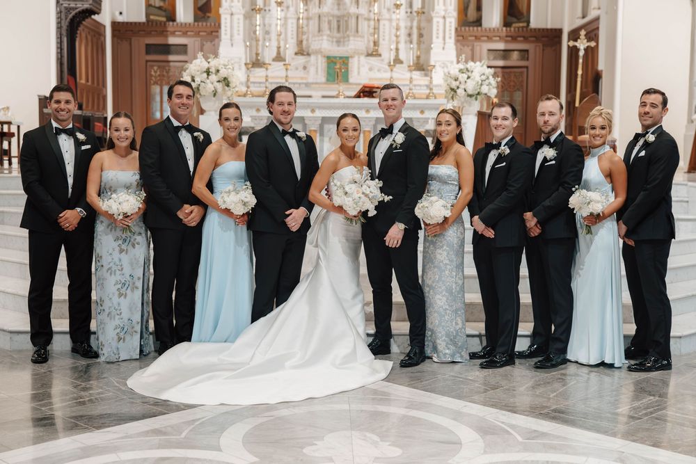 Wedding party poses inside elegant church with bridesmaids in light blue gowns and groomsmen in black tuxedos.