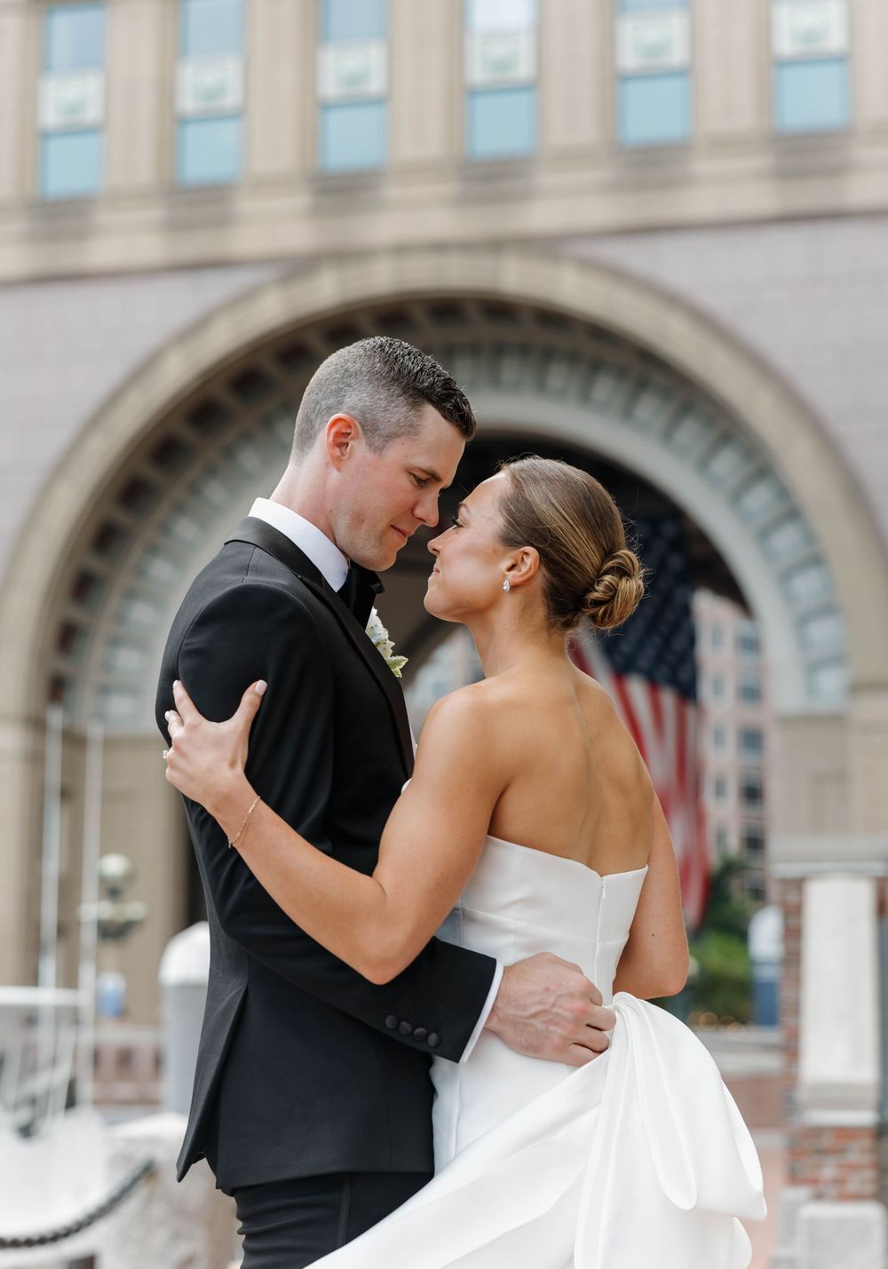 Bride and groom share a romantic embrace outside a grand building with an American flag backdrop on their wedding day.