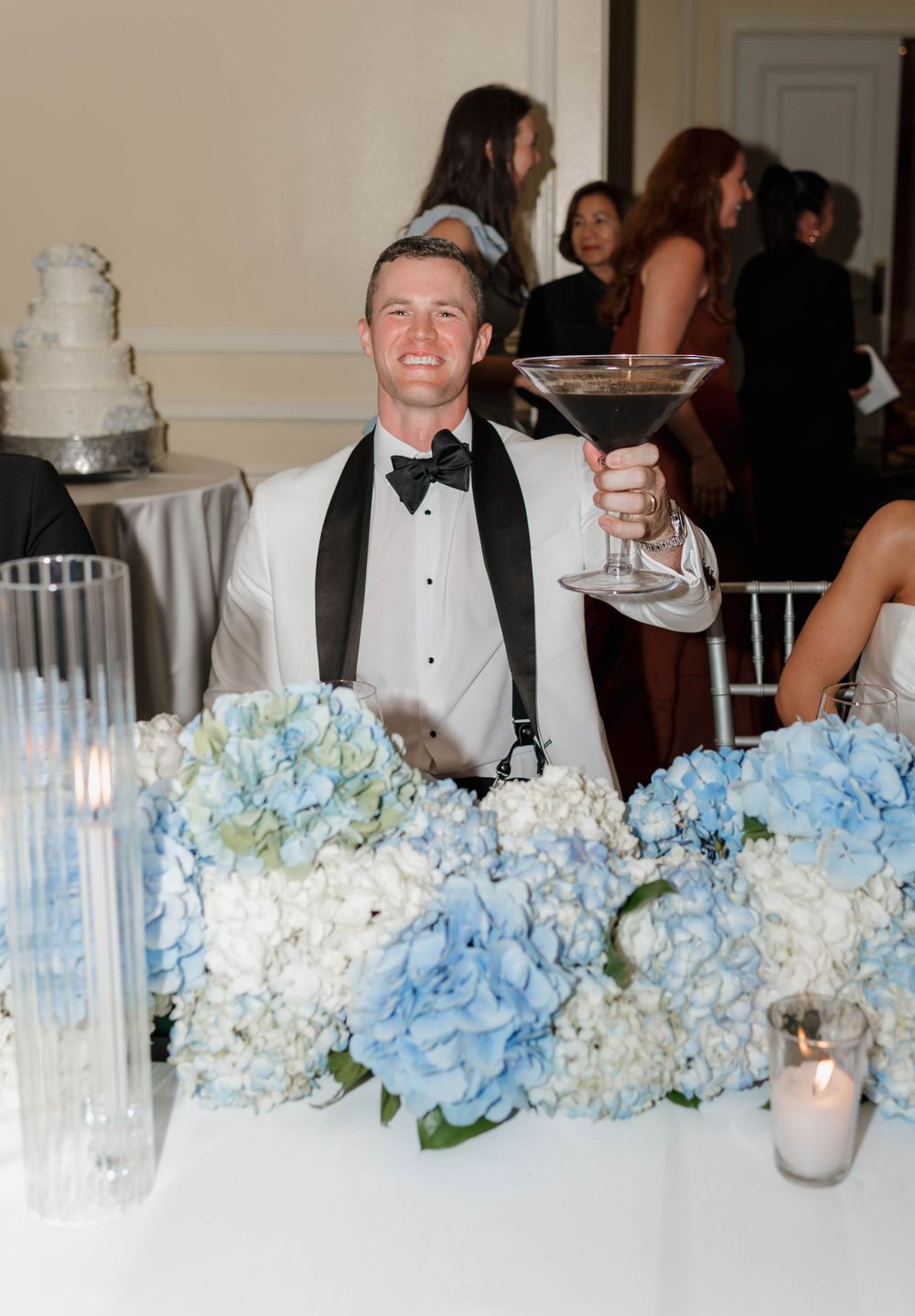 Smiling groom in white tuxedo holding oversized martini glass at wedding reception with blue hydrangea centerpieces.