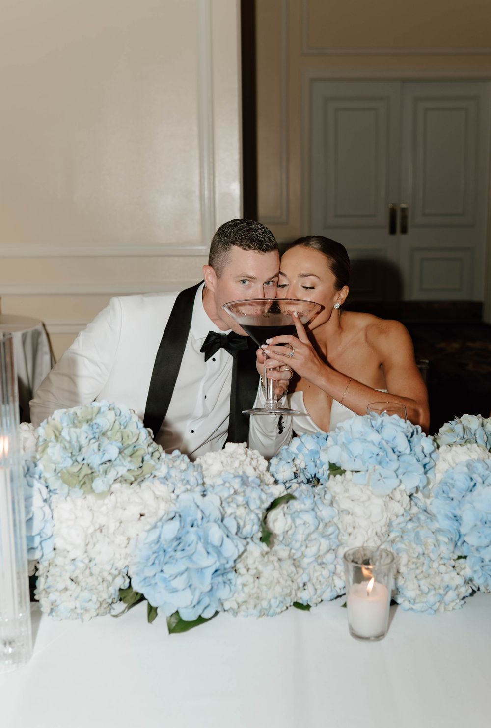 Bride and groom share a toast at their sweetheart table adorned with blue and white hydrangea floral arrangements.