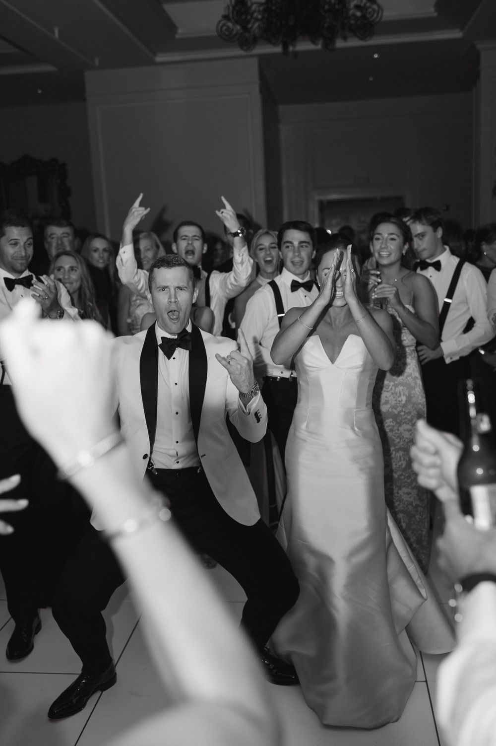 Bride and groom celebrate joyfully on the dance floor surrounded by energetic wedding guests in black and white photo.