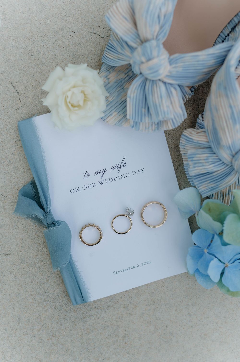 Wedding flat lay featuring three gold rings, a white rose, blue hydrangeas, and a vow book on a blue cover.