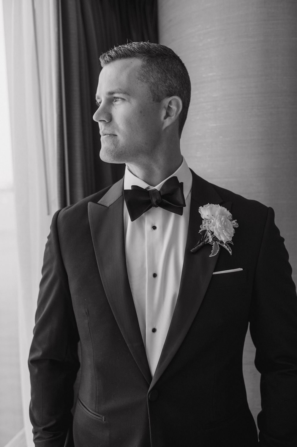 Elegant groom in black tuxedo with boutonniere gazing out window in classic black and white portrait.