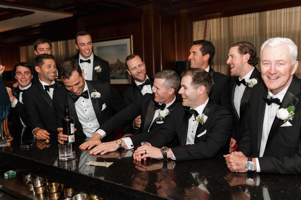 Groom and groomsmen in black tuxedos laugh together at a bar during wedding celebration.