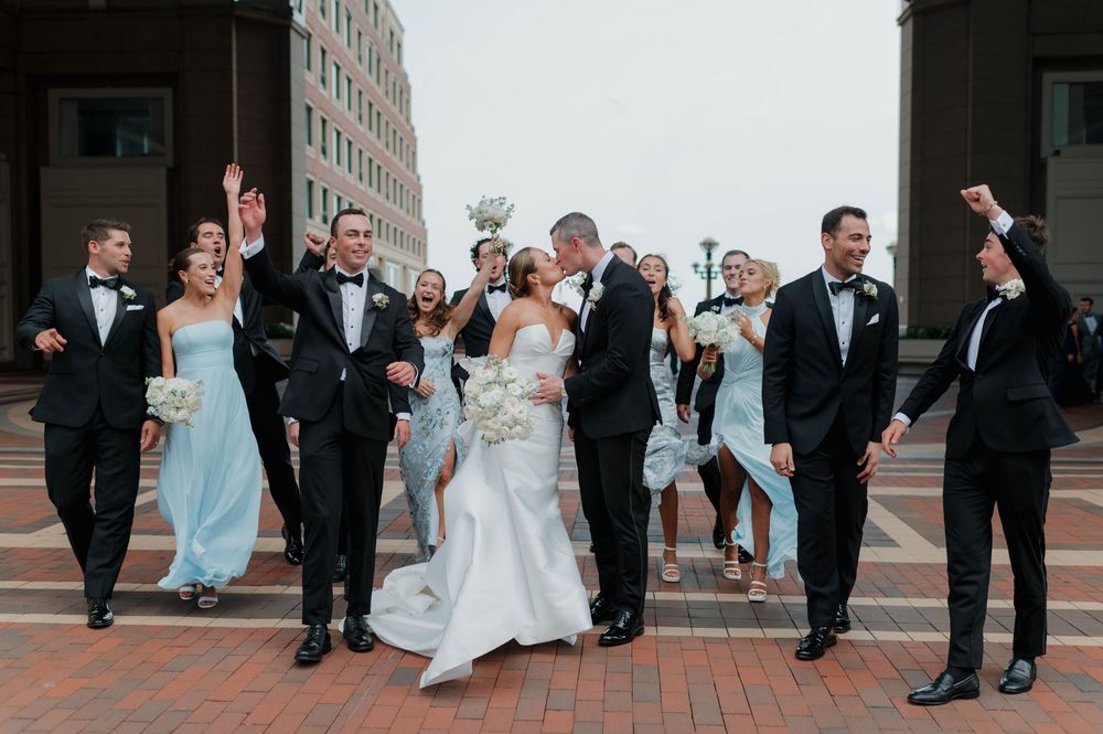Wedding party celebrates outdoors, bride and groom kiss while bridesmaids in blue and groomsmen in tuxedos cheer.