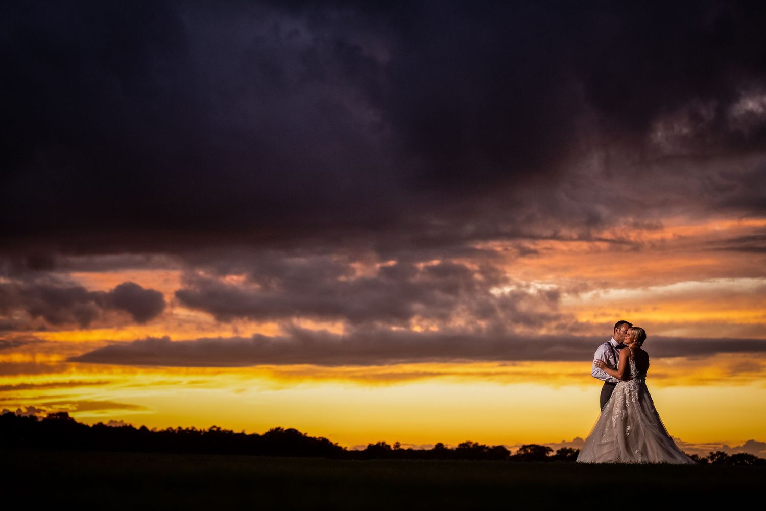Bride and groom embracing at sunset under dramatic storm clouds with vibrant orange and yellow sky.