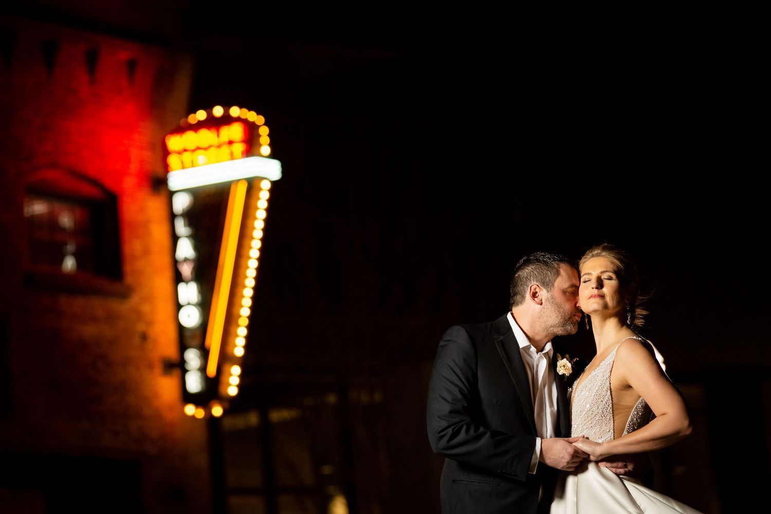 Bride and groom share a romantic kiss at night with glowing marquee lights behind them.