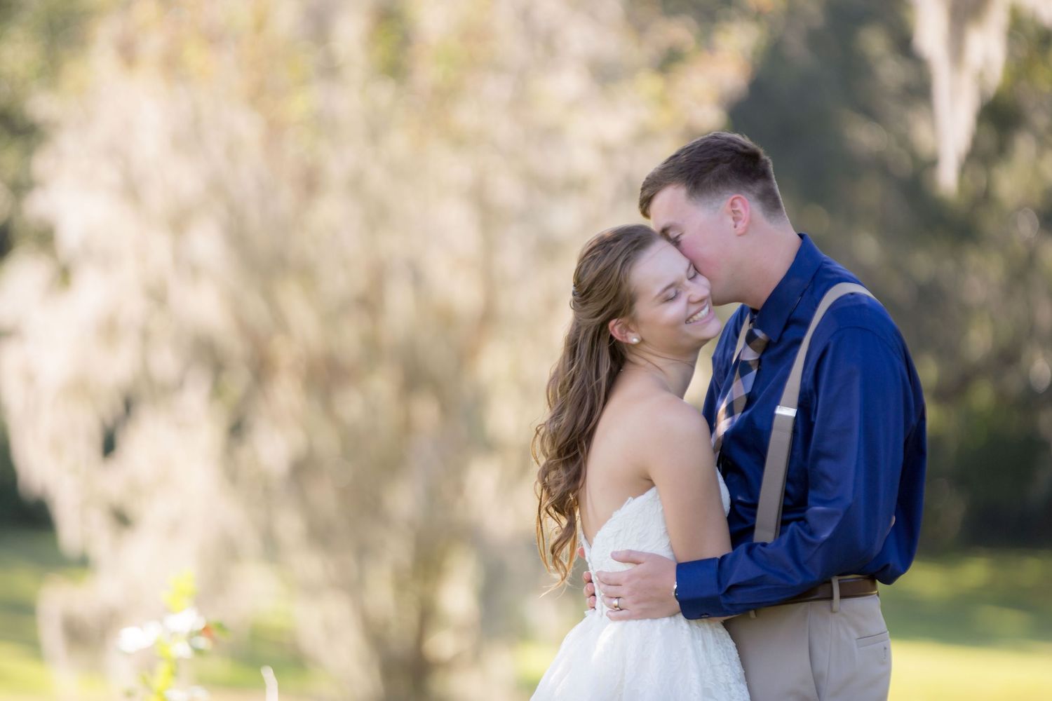 Bride and groom share a romantic embrace outdoors, with the groom in a blue shirt and suspenders nuzzling the smiling bride.
