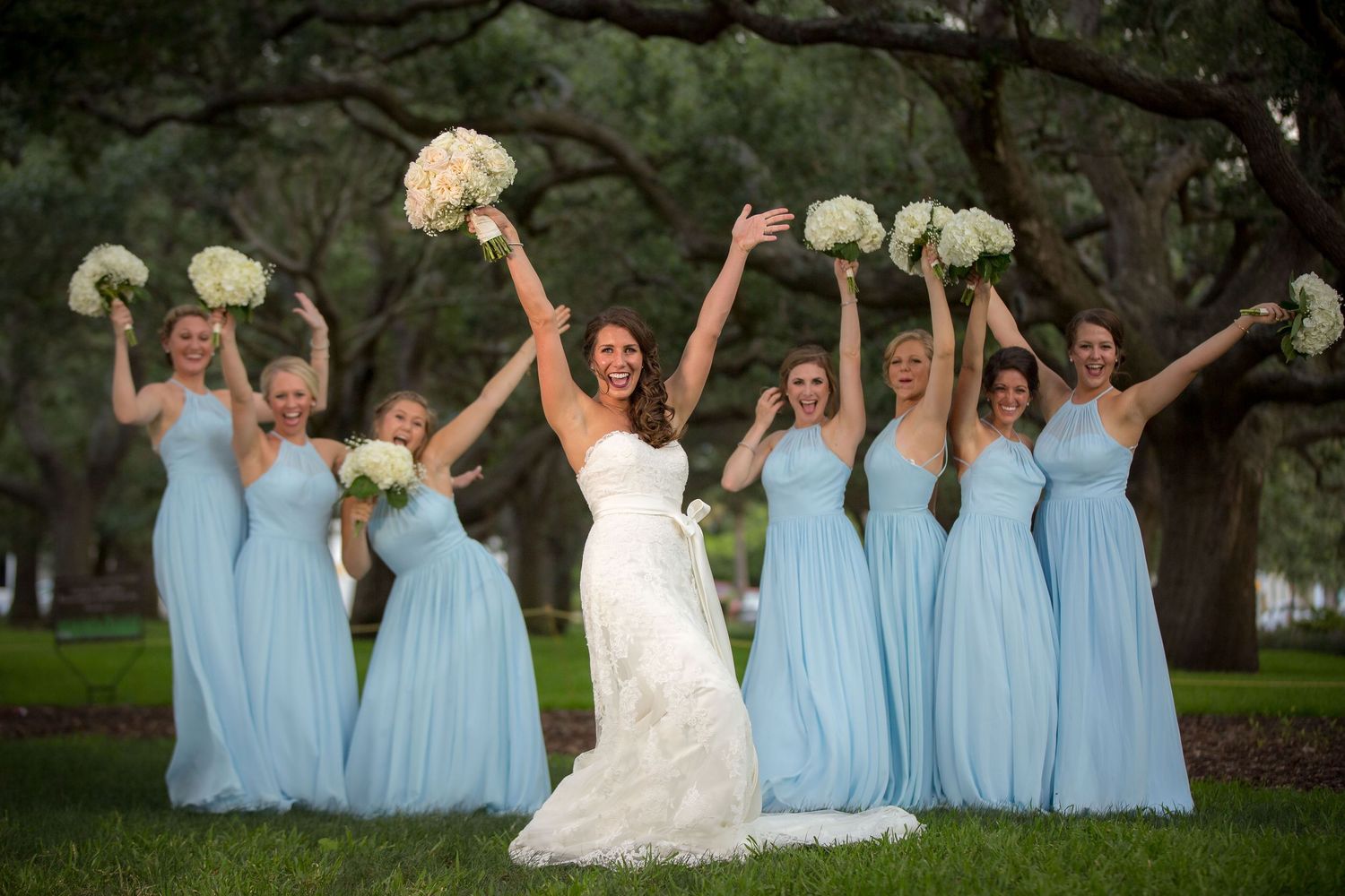Bride in white lace gown celebrates with bridesmaids in light blue dresses holding white bouquets outdoors under oak trees.