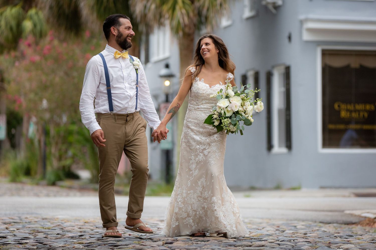 Bride and groom holding hands walking on cobblestone street, bride in lace gown holding white bouquet, groom in suspenders.