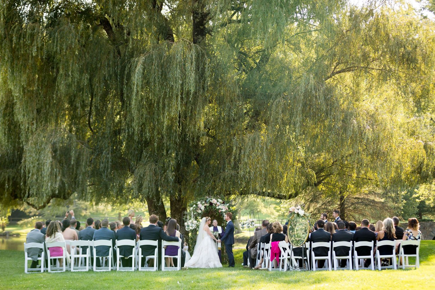 Outdoor wedding ceremony under a weeping willow tree with guests seated in white chairs on a lush green lawn.