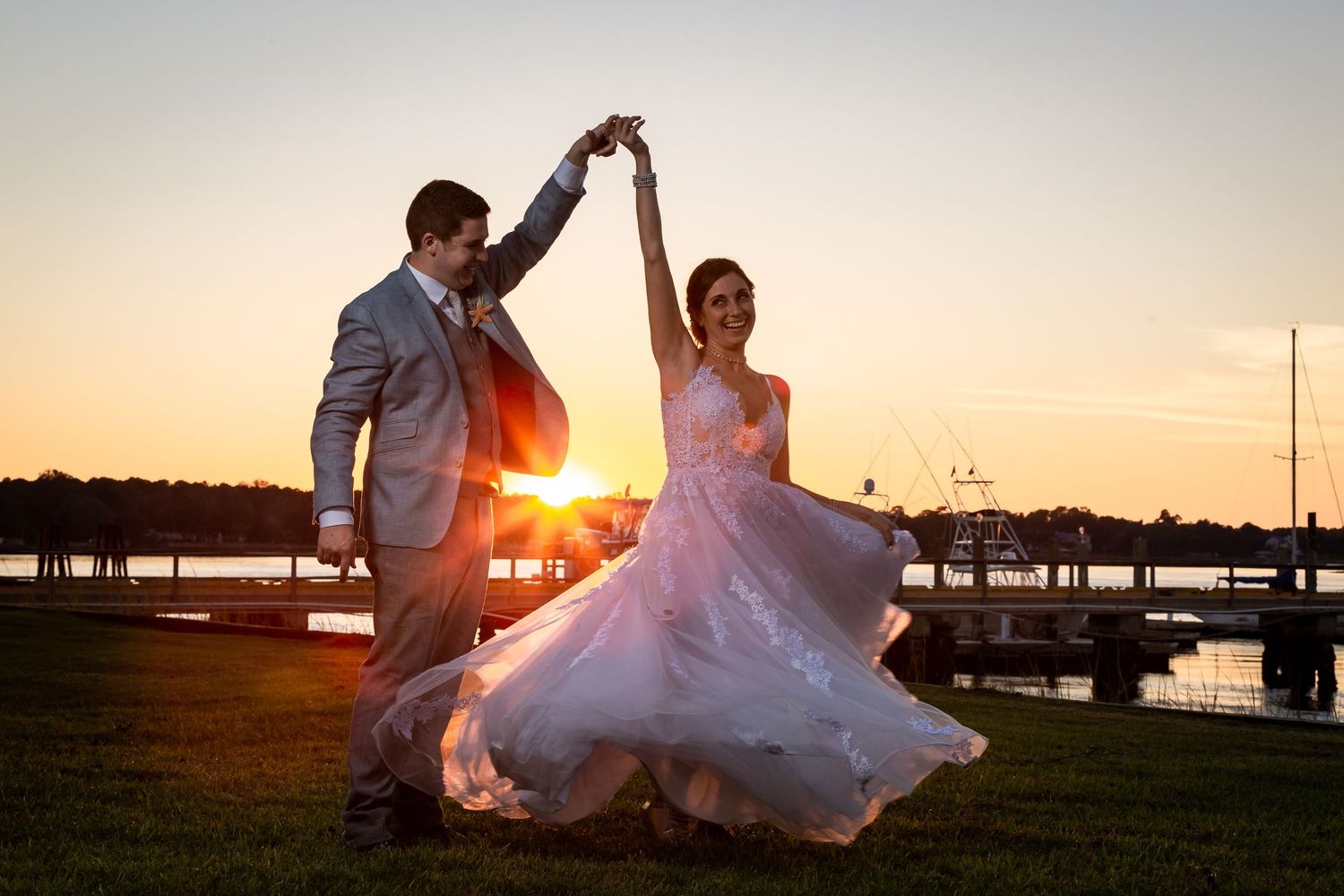 Bride and groom dancing at sunset by waterfront, wedding dress flowing, golden hour light.