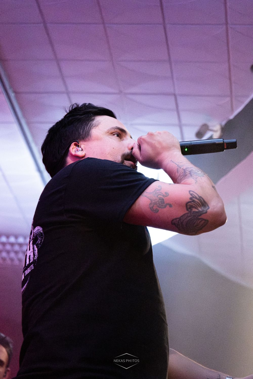 Tattooed male singer performing live on stage, holding microphone close to mouth, wearing black t-shirt under purple stage lighting.