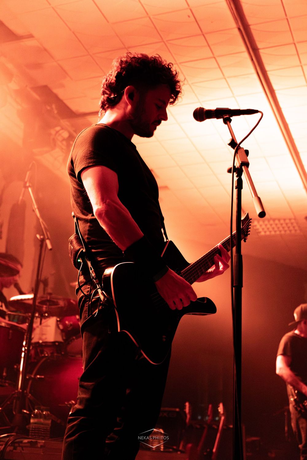 Male guitarist performing on stage under dramatic red lighting, playing electric guitar at a live rock concert.