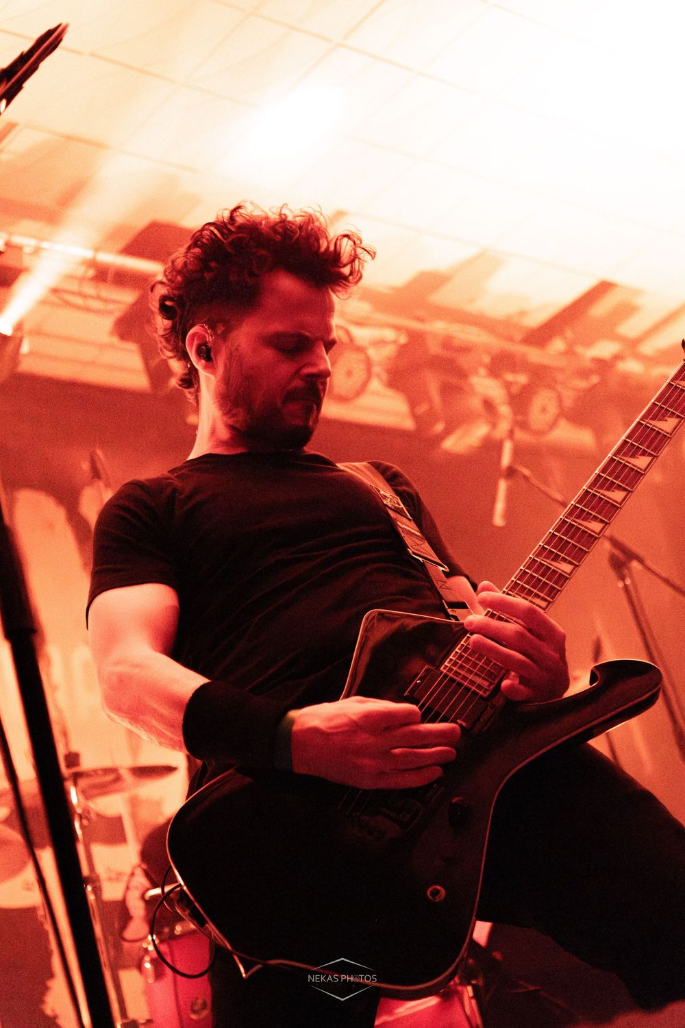 Male guitarist with curly hair plays electric guitar on stage under dramatic red lighting during a live concert performance.