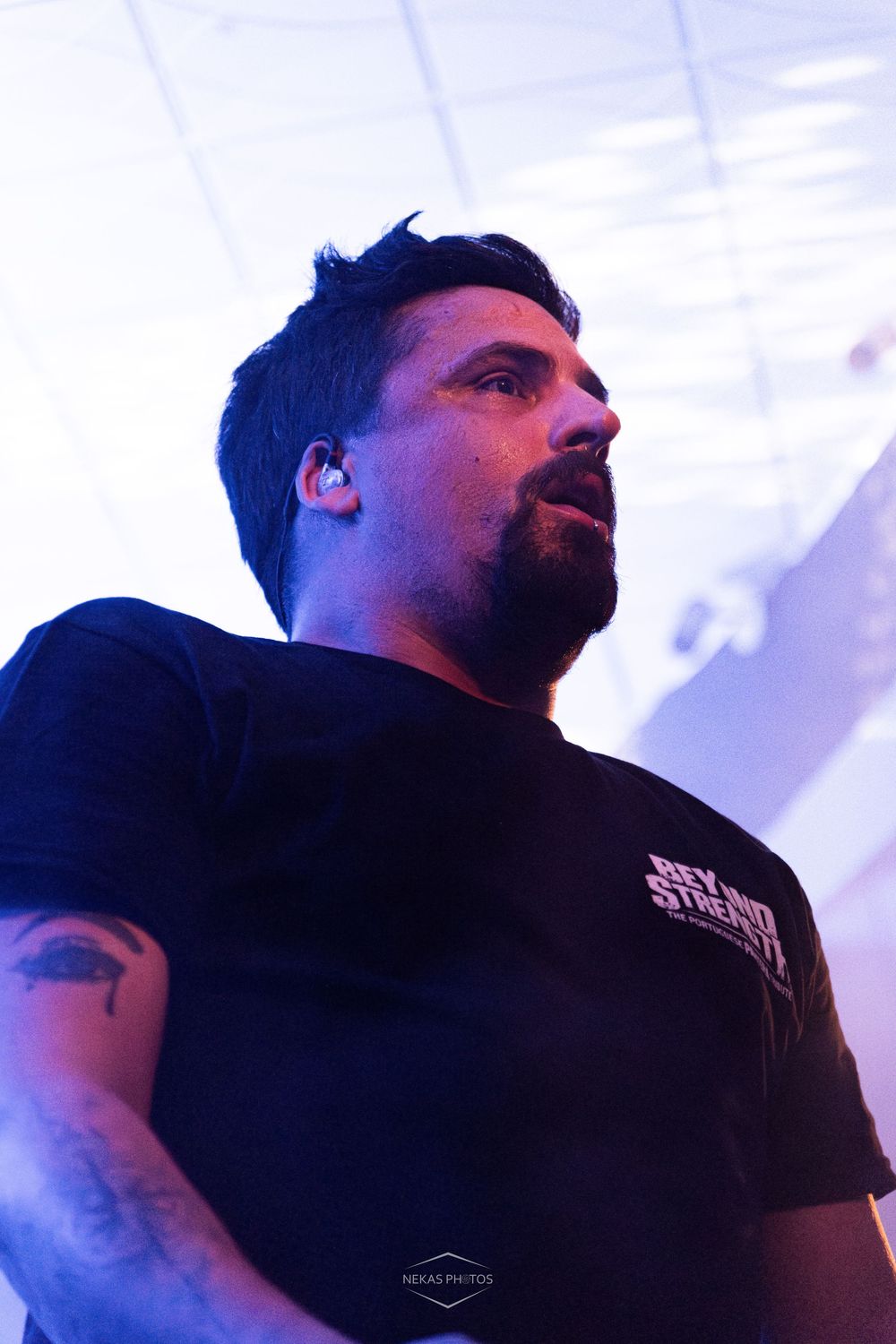 Man with beard and tattoos wearing black t-shirt performs on stage under blue and purple lights.