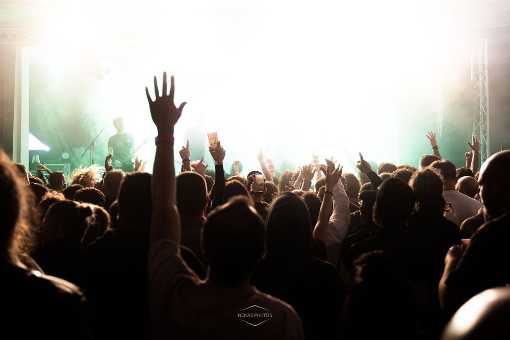Crowd of concert fans raising hands toward bright stage lights at a live music event.