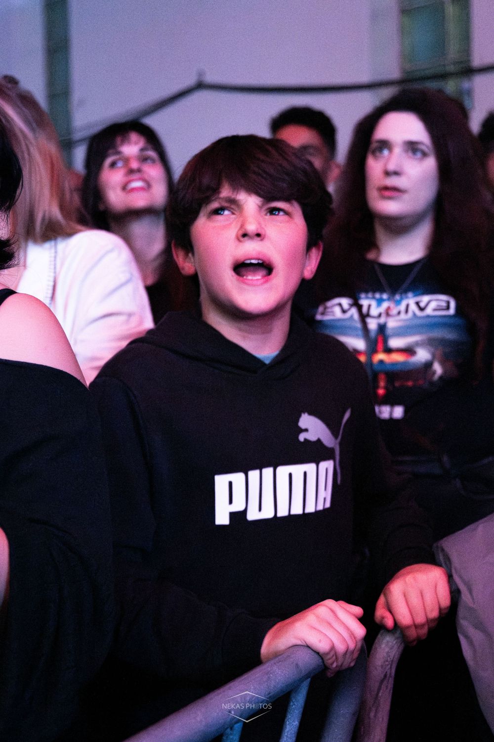 Young boy in black Puma hoodie with mouth open at a live concert, crowd visible behind him.