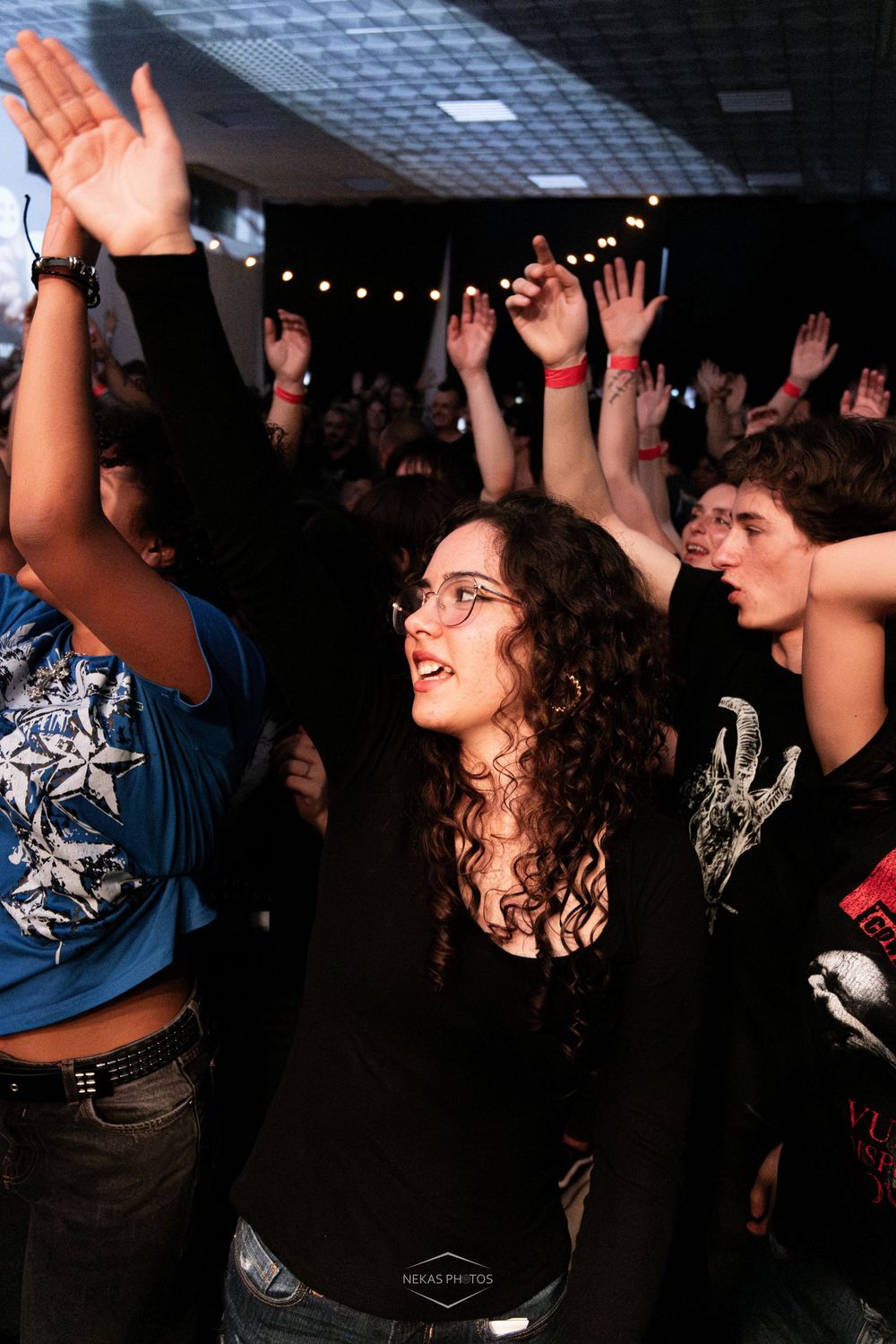 Excited concert crowd with hands raised, young woman with curly hair and glasses enjoying live music event indoors.