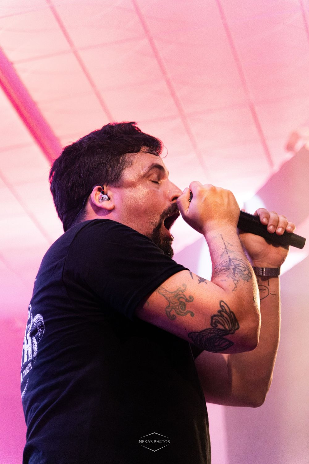Tattooed male vocalist singing passionately into microphone on stage under pink lighting at live concert.