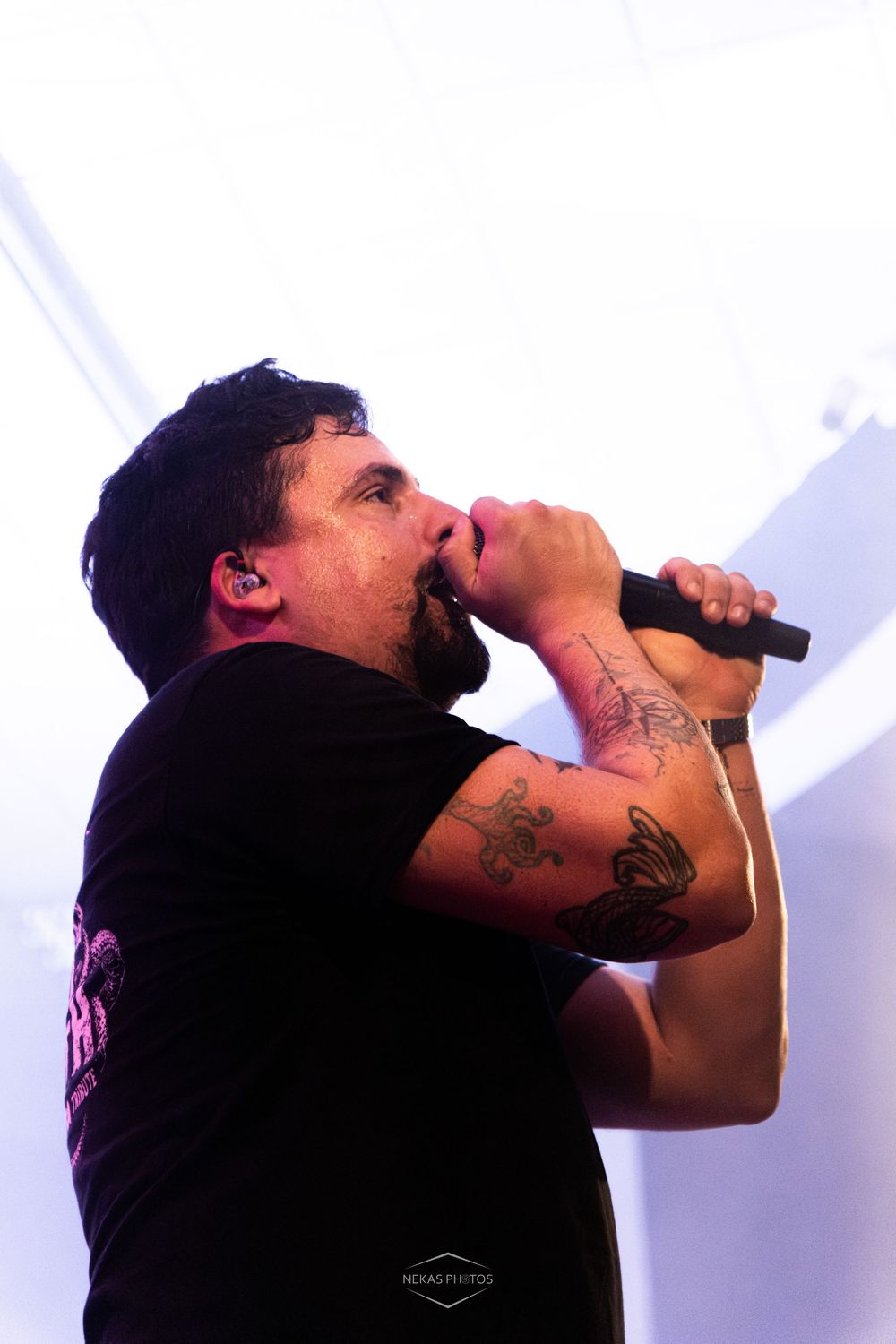 Tattooed male vocalist performing on stage, singing into a microphone under bright lights at a live concert.