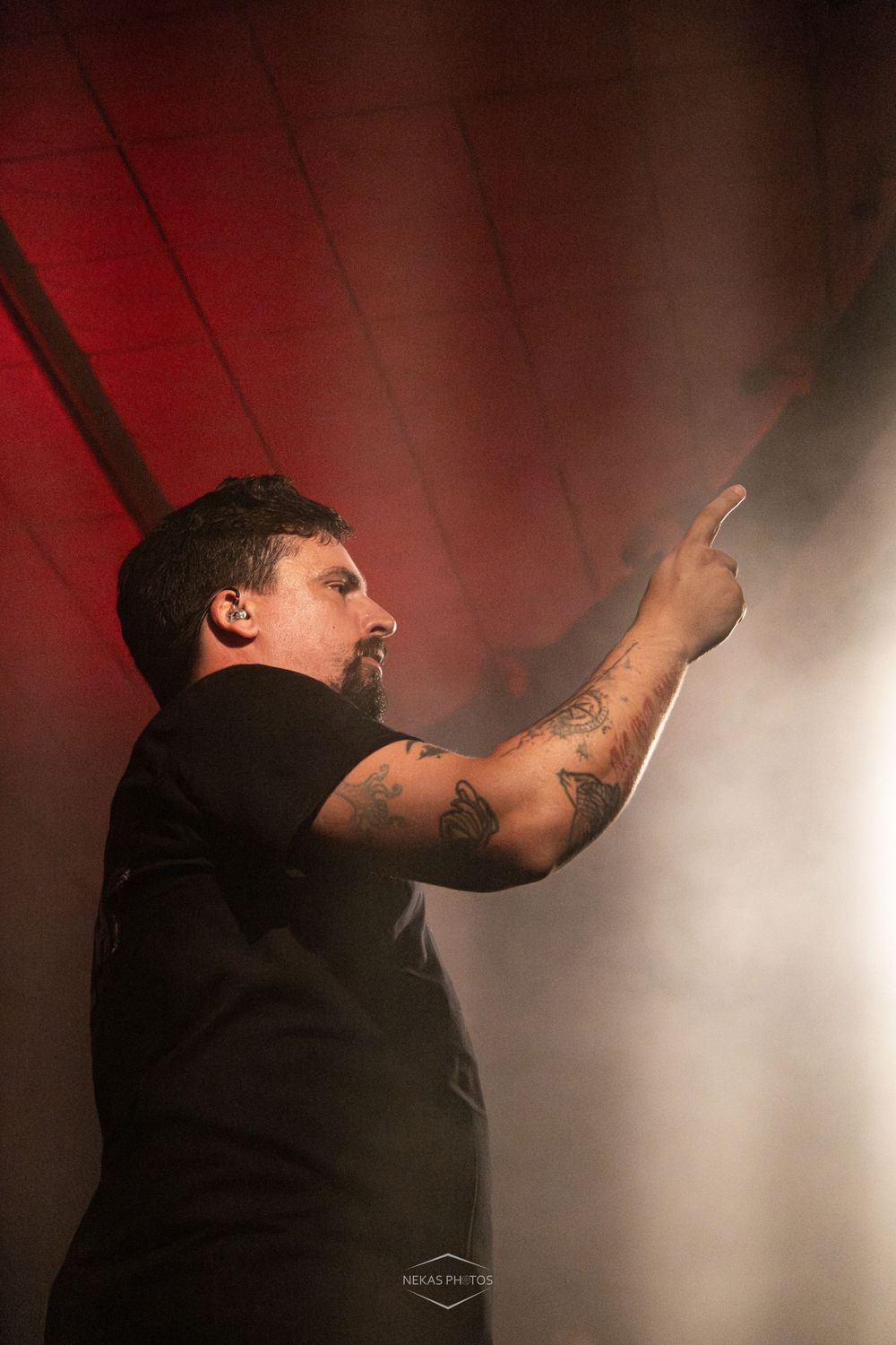 Tattooed male performer on stage pointing upward dramatically, surrounded by fog and red lighting effects.